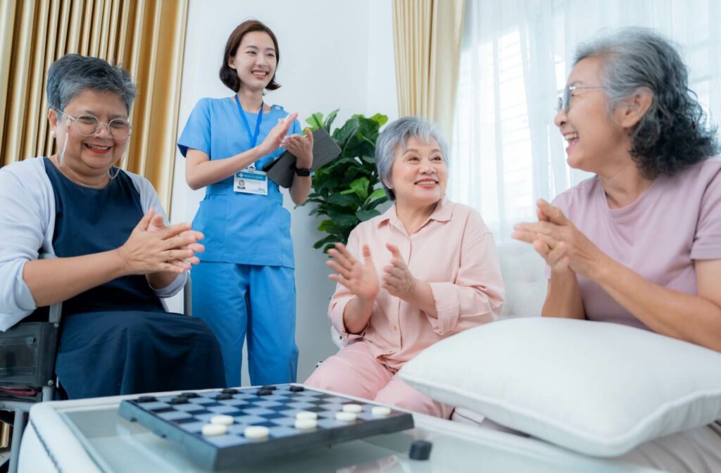 Group of older adults clapping and smiling with a caregiver in blue scrubs during a board game activity in a bright living room