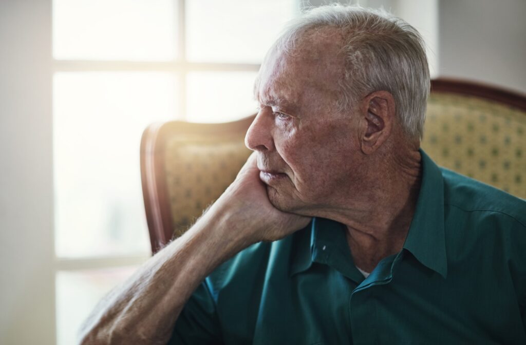 Older adult sitting alone in a chair looking out the window with a pensive expression in soft natural light.