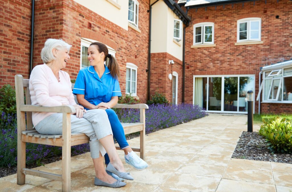 A resident in assisted living sits beside a smiling caregiver on an outdoor bench in a courtyard.