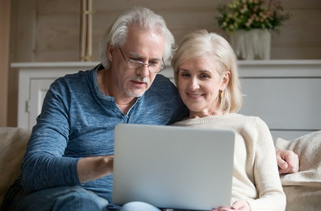A senior couple uses a laptop together while sitting on a sofa, looking at assisted living communities online.