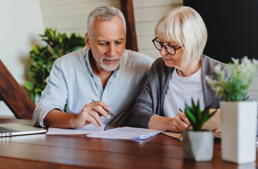 An older adult holding a pen sits beside their spouse at a desk and reviews paperwork to find out if they can afford assisted living together