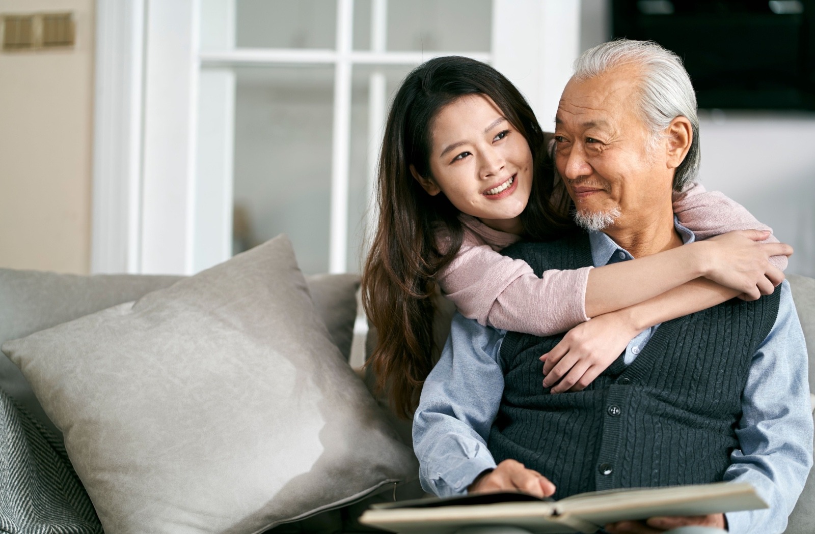 a younger adult smiles as they embrace their senior loved one who is sitting on a gray sofa
